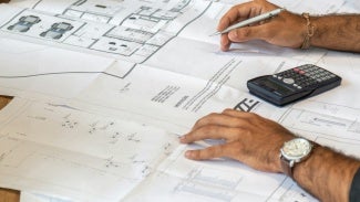 architectural drawings spread on a table with several people's hands, a calculator, and a laptop
