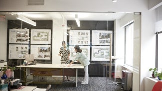 Two women collaborate at a table, reviewing design plans displayed on the walls, representing equity and accessibility in the architecture profession.